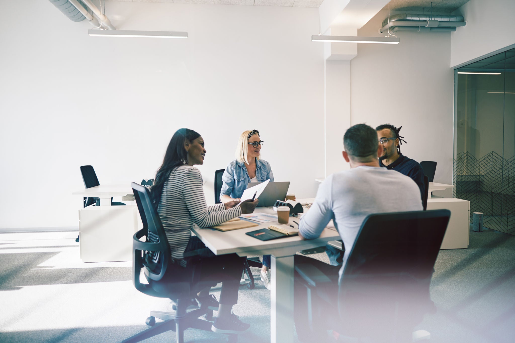 Smiling group of diverse coworkers talking together during a meeting around a table inside of a glass walled office