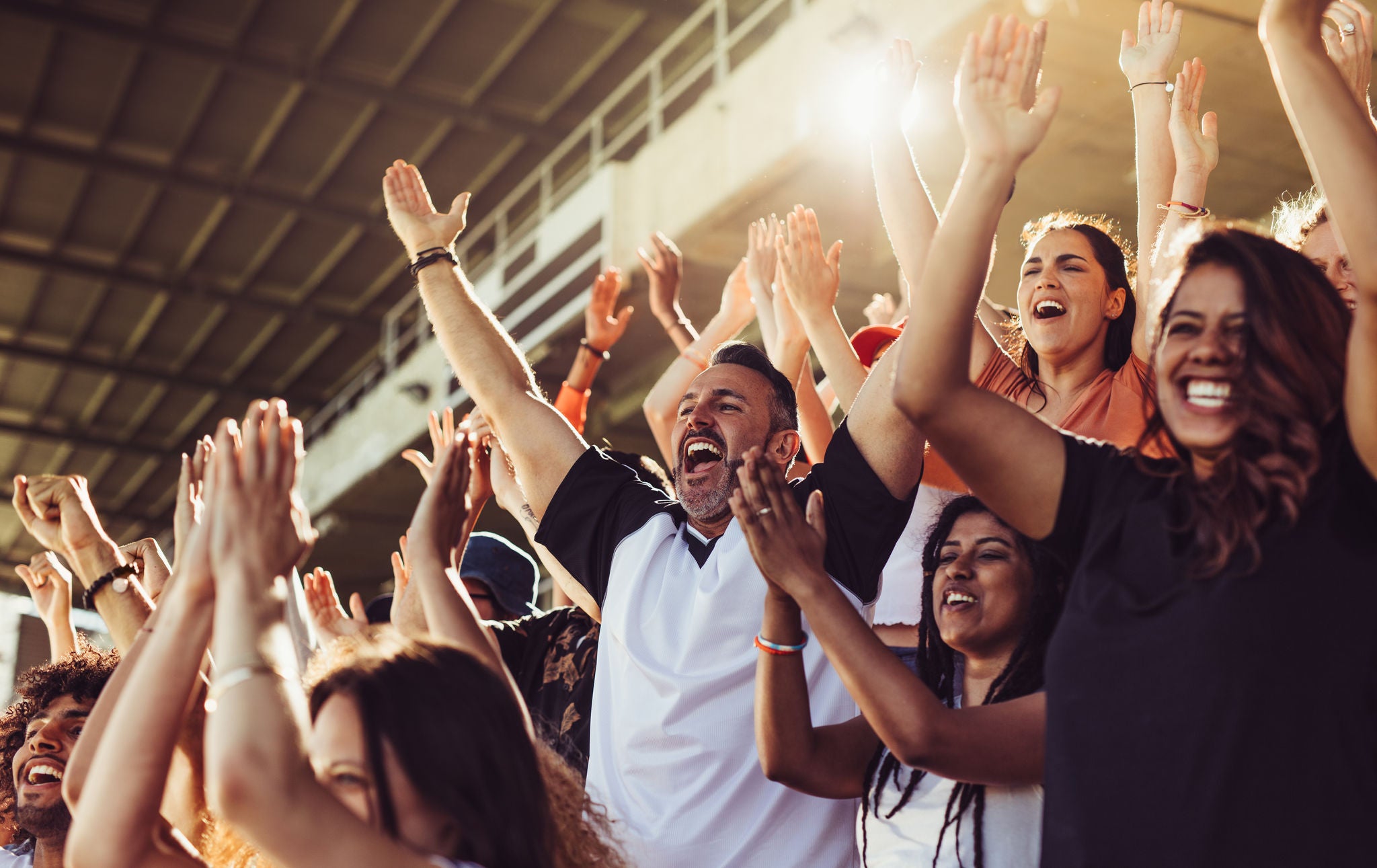 Crowd of sports fans cheering during a match in stadium. Excited people standing with their arms raised, clapping and yelling to encourage their team.