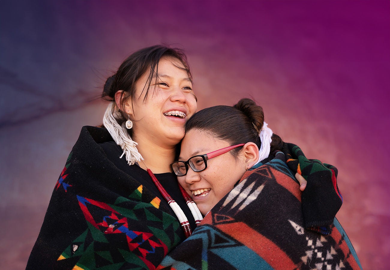 Two women hug while wearing Native American clothing.